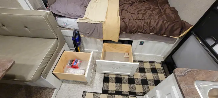 An RV rental's clever interior: open drawers beneath a bed and dinette reveal storage space, with a small vacuum tucked away. Features a checkered rug.. Forest River Rockwood Geo Pro 2022