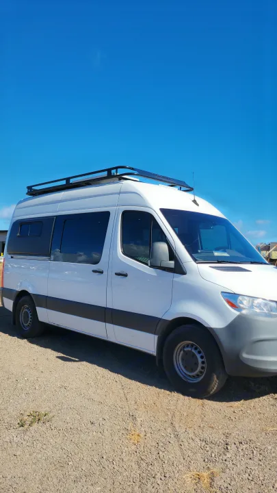White Mercedes-Benz Sprinter van conversion with a roof rack, ready for your RV rental adventure. Parked on gravel.. Mercedes-Benz Sprinter 2021