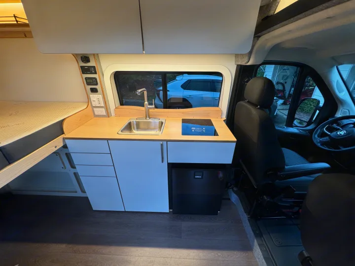 This modern RV rental interior showcases a fully equipped kitchenette with a sink, cooktop, and fridge, plus a comfortable bed and control panel.. Dave & Matt Vans Dave & Matt Vans Class B 2024