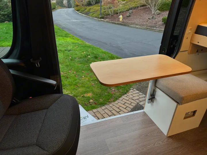 RV rental interior shows a light wood table, cushioned bench, and driver's seat with a view of a road and grass outside.. Dave & Matt Vans Dave & Matt Vans Class B 2024