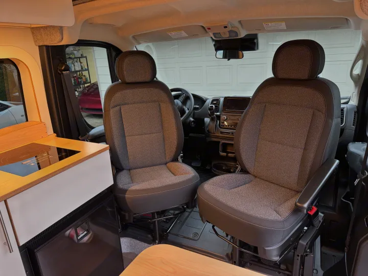 Explore an RV rental's cozy interior: front seats, dashboard, and a compact kitchenette with induction cooktop and fridge.. Dave & Matt Vans Dave & Matt Vans Class B 2024