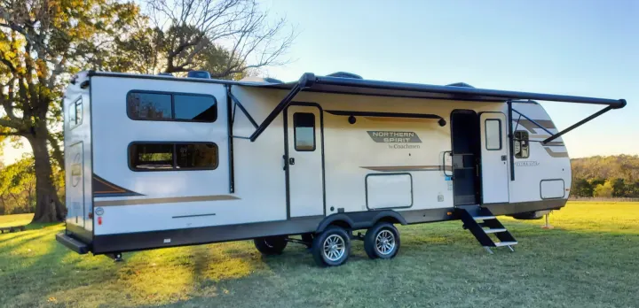 White & tan Northern Spirit travel trailer, an RV rental, parked on grass with awning extended and steps down.. Coachmen Northern Spirit 2024