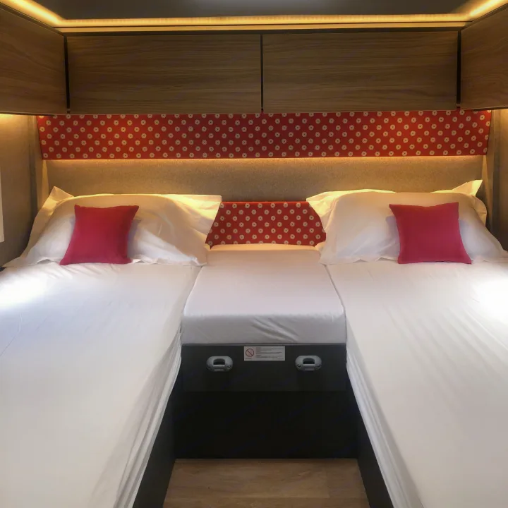 Twin beds in a compact RV rental interior, with a vibrant red patterned headboard, overhead cabinets, and soft lighting.. GlamperRV DBL 2026