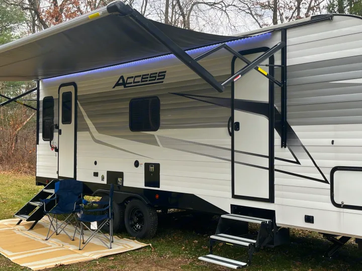 White RV rental travel trailer with extended awning, blue LEDs, and outdoor chairs set up for camping.. Winnebago Access 26 RL 2024