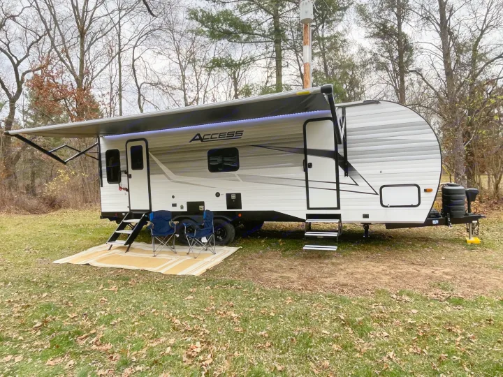 This RV rental, an 'Access' travel trailer, is set up at a woodsy campsite with an extended awning and chairs.. Winnebago Access 26 RL 2024