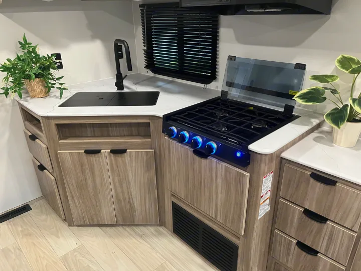 Compact RV rental kitchen with light wood cabinets, white countertops, black sink, and a modern blue-lit gas stove.. Winnebago Access 26 RL 2024