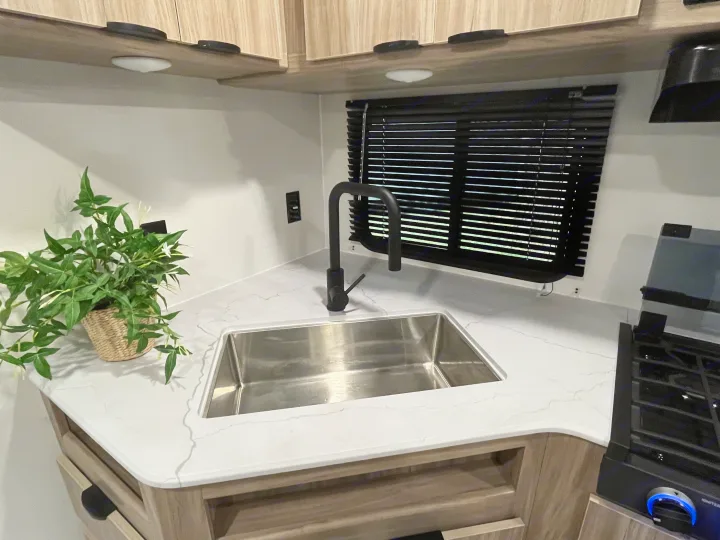 Modern RV rental kitchen: white counters, black faucet, stainless sink, compact stove, and wood-look overhead cabinets.. Winnebago Access 26 RL 2024