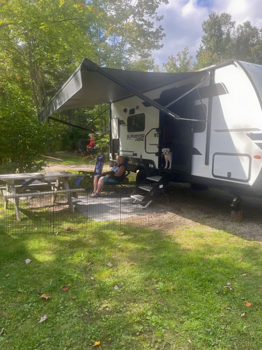 Relaxing at an RV rental campsite. A person, dog, and picnic table are outside a white travel trailer with an awning.. Forest River Surveyor Legend 19SSLE 2024