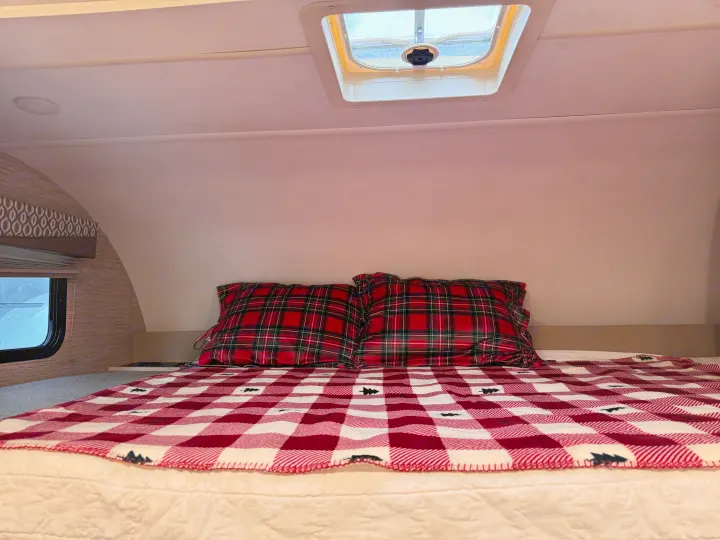 Inside a cozy RV rental: a comfortable bed with festive plaid pillows & a tree-patterned blanket. Window shows snow.. Thor Freedom Elite 2022