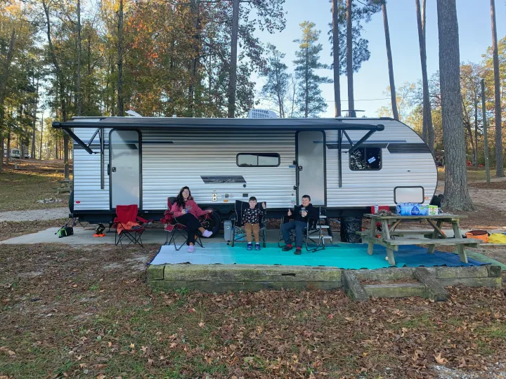 Family enjoying their RV rental: A silver travel trailer with an awning extended, parked at a sunny campground.. Forest River East to West 2025