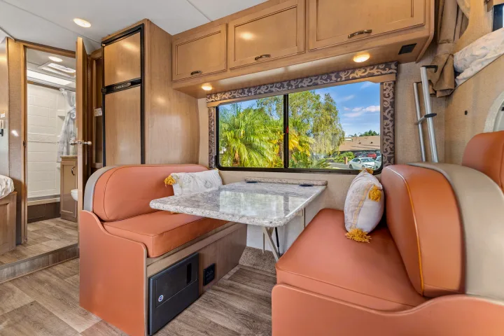 RV rental interior: inviting dinette with cushioned benches & table, compact kitchen with fridge, and a bright bathroom beyond.. Thor Four Winds 2019