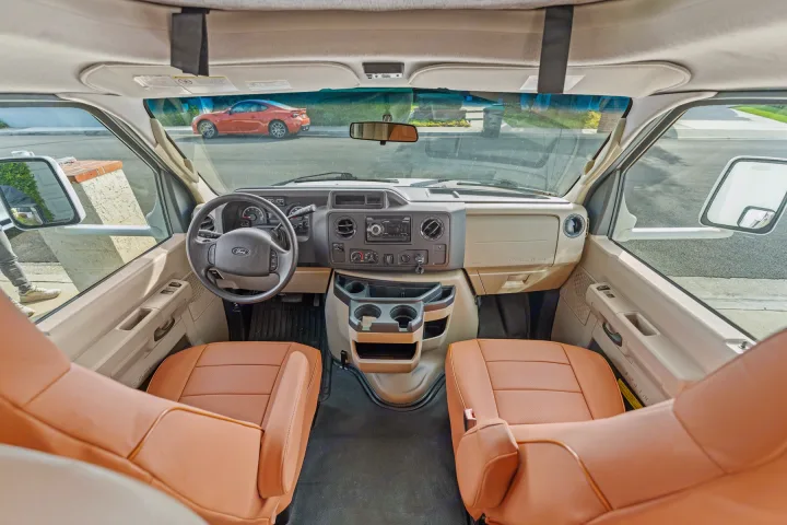 RV rental interior: wide view from the rear of the cabin showing the dashboard, steering wheel, tan front seats, and street outside.. Thor Four Winds 2019