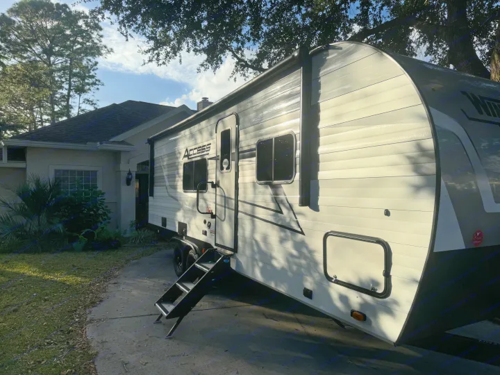 A bright white Access travel trailer rv rental, parked on a sunlit driveway next to a house, with steps extended.. Winnebago Access 2024
