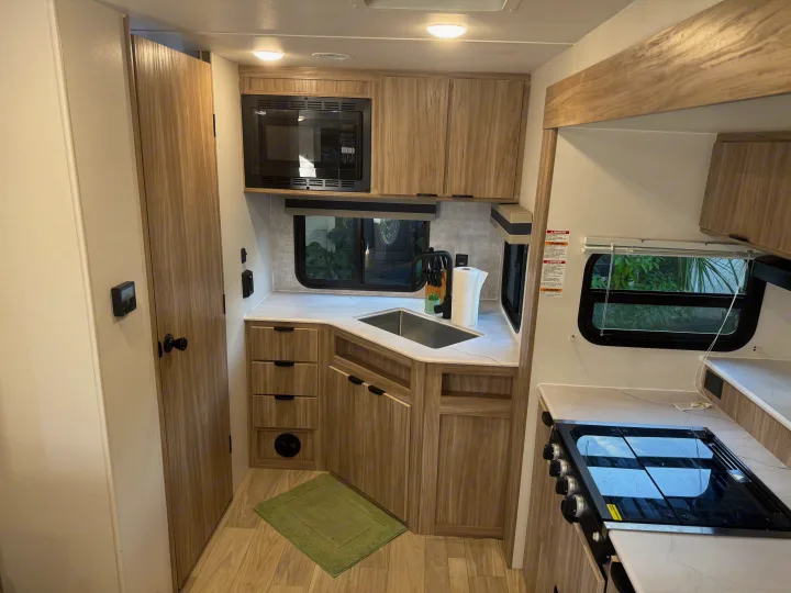 Modern RV rental kitchen featuring a microwave, sink, stovetop, and light wood cabinetry, with two windows.. Winnebago Access 2024