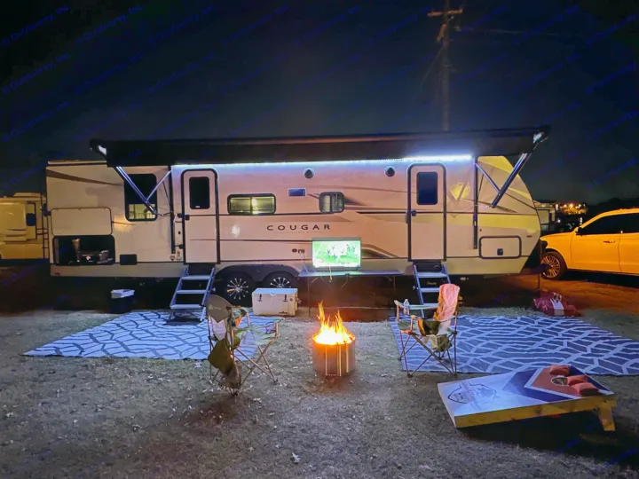 This RV rental offers ultimate outdoor comfort at night, featuring an illuminated awning, TV, fire pit, chairs, and cornhole game.. Keystone RV Cougar 2024