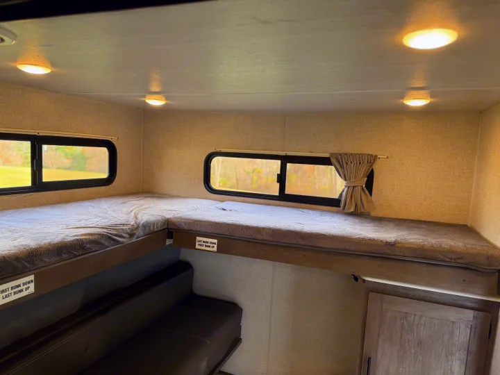 Cozy bunk beds in an RV rental interior, featuring a lower sofa bunk and large windows overlooking scenery.. Forest River Wildwood 2021