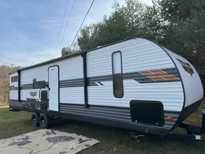 Modern white & grey travel trailer RV rental, with a mat on grass. Parked outdoors under a blue sky near trees.. Forest River Wildwood 2021