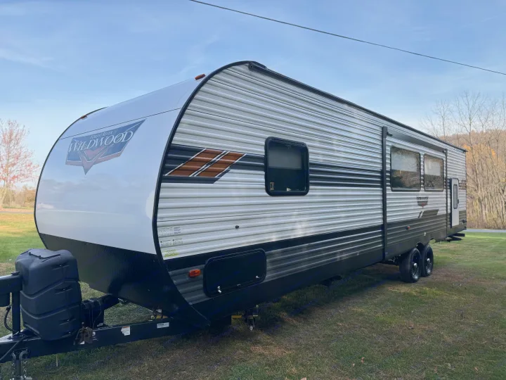 White and grey Forest River Wildwood travel trailer with brown accents, ideal for an RV rental, parked on grass under a clear sky.. Forest River Wildwood 2021