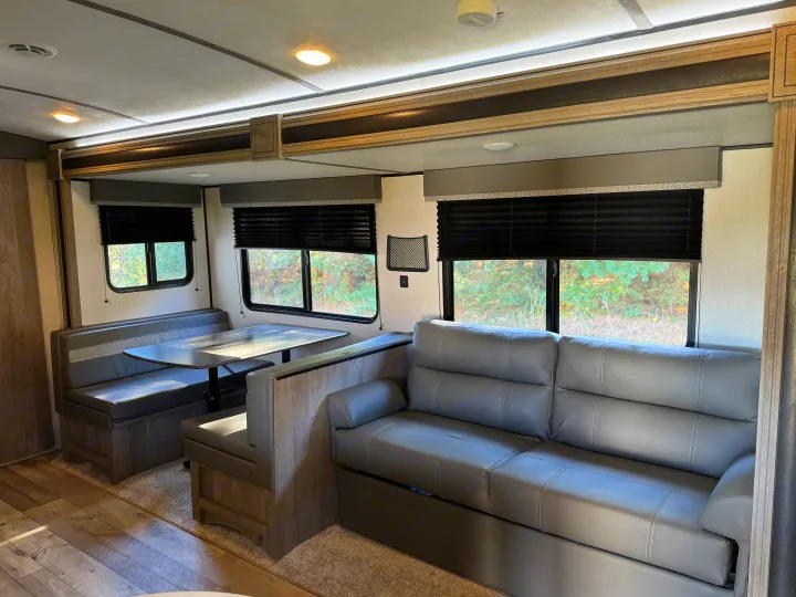 This rv rental shows a comfortable living area with a grey sofa, booth dinette, and large windows looking out to trees.. Keystone RV Springdale 2021