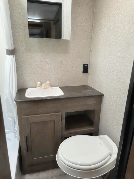 Compact **RV rental** bathroom. Features a light wood-look vanity, white sink, toilet, shower with curtain, and mirrored cabinet.. Keystone RV Springdale 2021