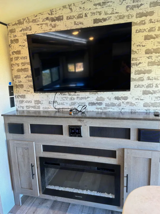 RV rental living area: A large TV above a media console with an electric fireplace & speakers, set against a stylish brick wall.. Keystone RV Springdale 2021