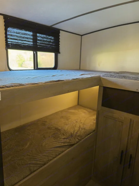 RV rental interior: bunk beds with gray blankets, a window with dark blinds, and cabinetry. Lower bunk is dimly lit.. Keystone RV Springdale 2021