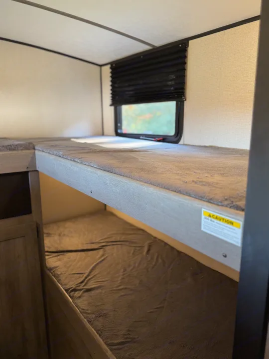 Cozy rv rental interior featuring bunk beds with soft grey blankets, a window, and a black pleated blind in a camper.. Keystone RV Springdale 2021