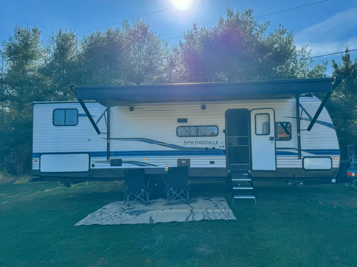 Sunny RV rental setup: a white and blue Springdale travel trailer with an extended awning, outdoor rug, and chairs on green grass.. Keystone RV Springdale 2021