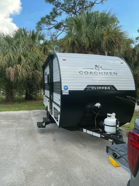 A Coachmen Clipper rv rental travel trailer is parked on a paved lot with palm trees. Propane tank & leveling block visible.. Coachmen Clipper Cadet 17CBH 2025