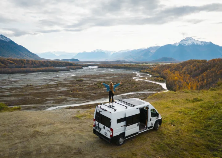 Scenic RV rental adventure: A person stands atop a van overlooking a vast river valley and majestic mountains.. Dodge Van 2024