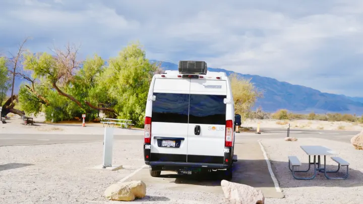 White RV rental (Winnebago Solis campervan) parked at a gravel campsite, surrounded by trees with mountains visible.. Winnebago Solis 59px 2025