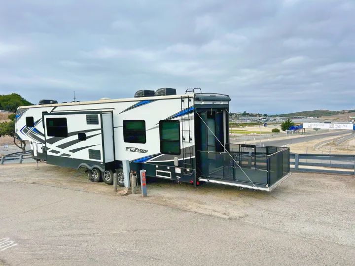 A white Fusion toy hauler RV rental with a patio deck overlooks a race track. Perfect for trackside camping!. Keystone Fuzion 2022