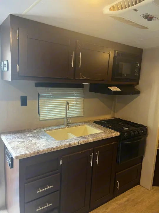 Modern RV rental kitchen featuring dark cabinets, light speckled countertops, a double sink, stove, microwave, and ceiling AC.. K-Z Manufacturing Sportsmen 2019