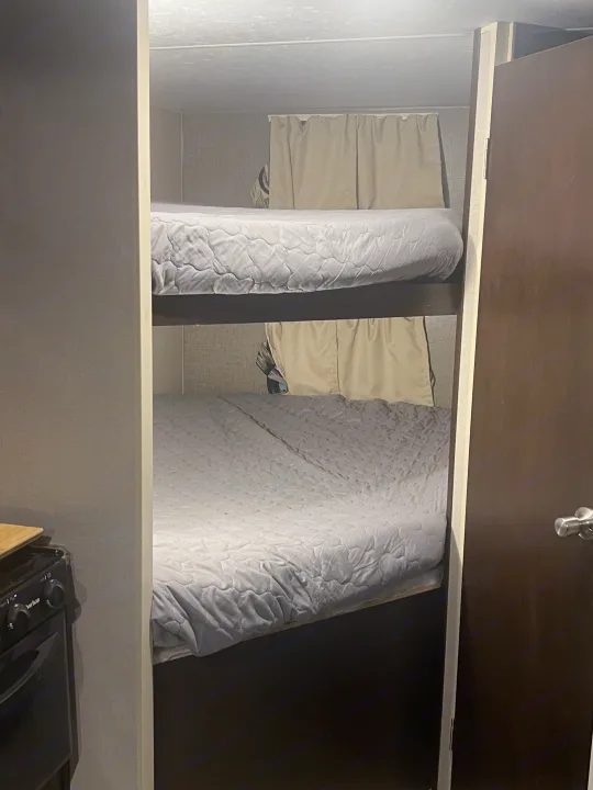 Compact RV rental interior featuring two bunk beds with grey bedding, a small kitchen unit, and a side door.. K-Z Manufacturing Sportsmen 2019