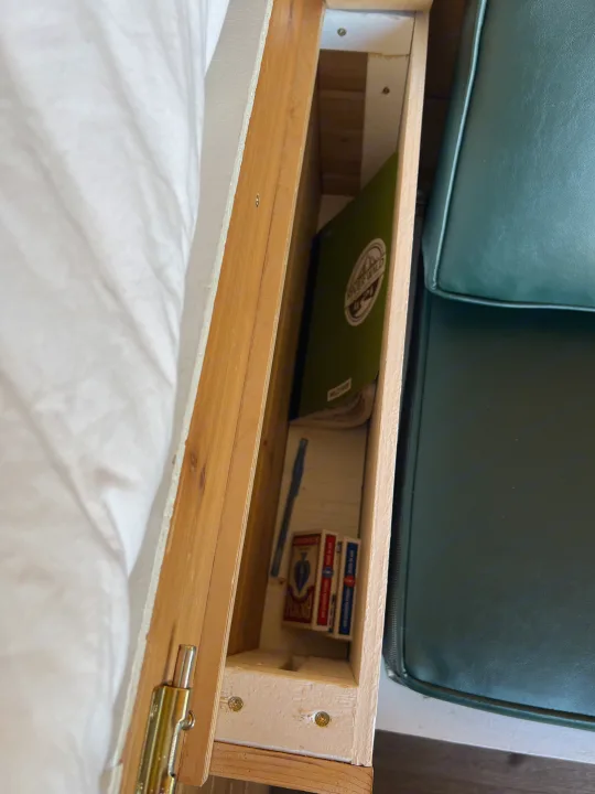 This image shows a narrow storage compartment in an RV rental, holding a notebook, pen, and playing cards for travel essentials.. Dodge Van 2024