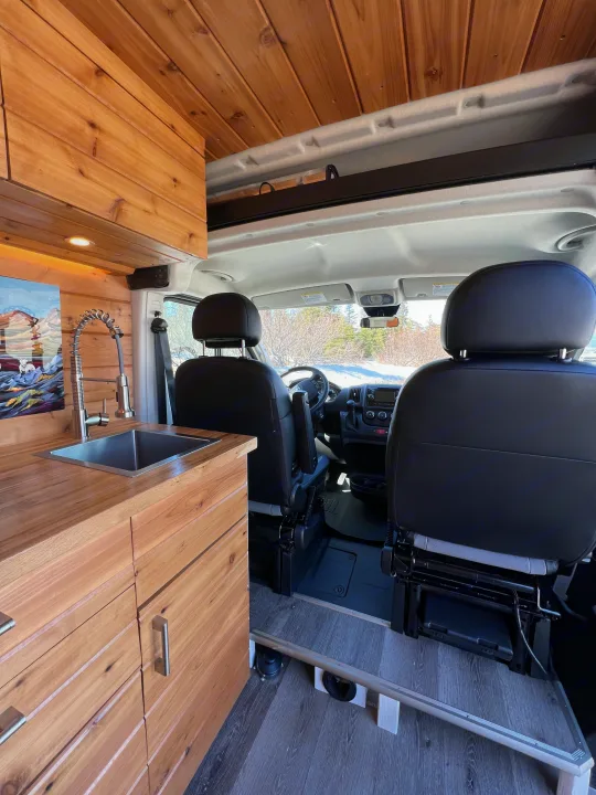 Inside this modern RV rental: a warm wood kitchen with sink on the left, leading to driver/passenger seats & snowy view.. Dodge Van 2024