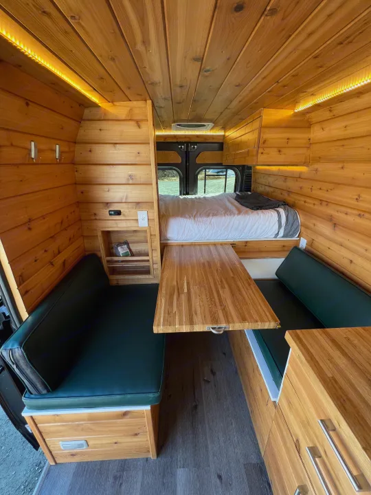 Cozy wood-paneled RV rental with a comfy bed, green-cushioned dinette, and custom storage for your next adventure.. Dodge Van 2024