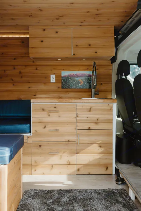 Inside an RV rental: a warm, wood-finished camper van interior with a kitchen, comfy blue seating, and ample storage.. Dodge Van 2024