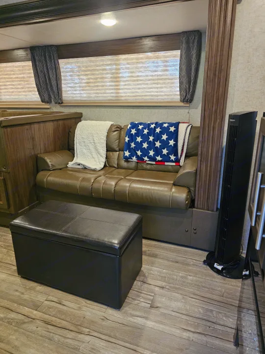 RV rental interior: cozy living area with a brown sofa, patriotic blanket, black ottoman, and tower fan.. Forest River Cherokee Grey Wolf 2019