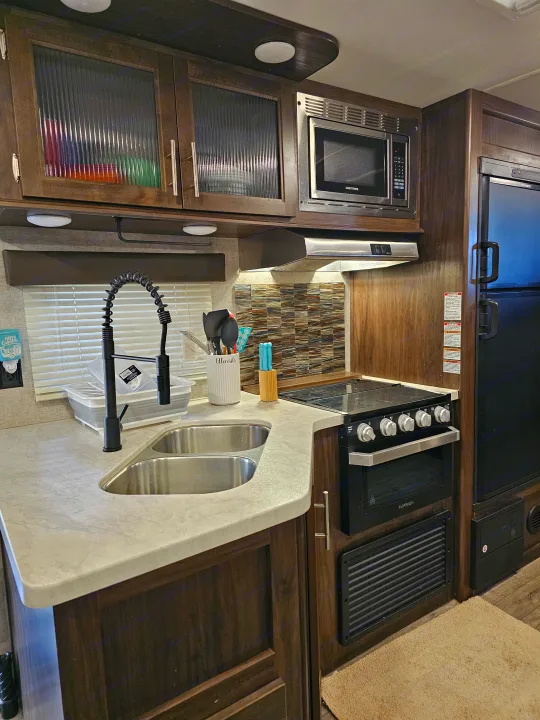 An RV rental kitchen with modern dark wood cabinets, a stainless steel microwave, stove/oven, double sink, and tiled backsplash.. Forest River Cherokee Grey Wolf 2019