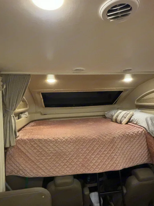 Inside an RV rental: a spacious cab-over bed with a soft, quilted cover and pillows, illuminated by ceiling lights.. Entegra Coach Odyssey 2021