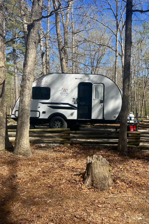 A 'Joy Ride' RV rental, a white travel trailer, settled in a peaceful, wooded campsite with a log fence and generator.. Braxton Creek Bushwhacker Plus 2023
