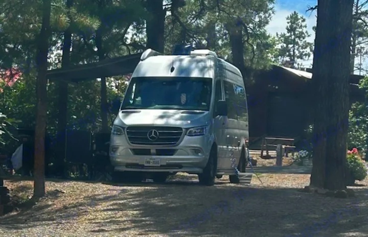 A silver Mercedes-Benz Sprinter RV rental is parked among tall trees in a sunny campground, offering a shaded outdoor experience.. Airstream Interstate 2020