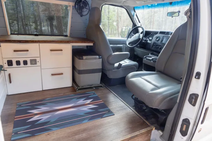 RV rental interior: custom wood cabinets, pull-out fridge, grey seats, and a forest view from the side window of this camper van.. Ford Econoline 150 2009