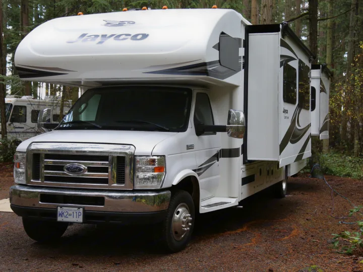 A white Jayco motorhome, an RV rental, parked amidst green trees with slide-outs extended for extra space.. Jayco Greyhawk 2019