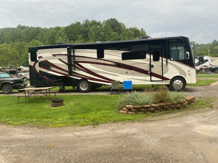 Large Encore RV, like an rv rental, parked on a gravel site at a campground with a picnic table, fire pit, and chairs. Forest background.. Coachmen Encore 2022