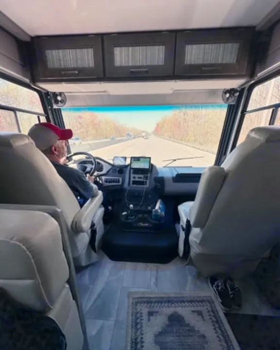 Inside an RV rental, a man drives on a highway. This motorhome interior view shows the cockpit, passenger seat, and road ahead.. Coachmen Encore 2022