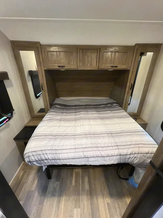 Cozy RV rental bedroom featuring a striped bed, wood-look overhead cabinets, and mirrored bedside storage.. Forest River Vibe 2020