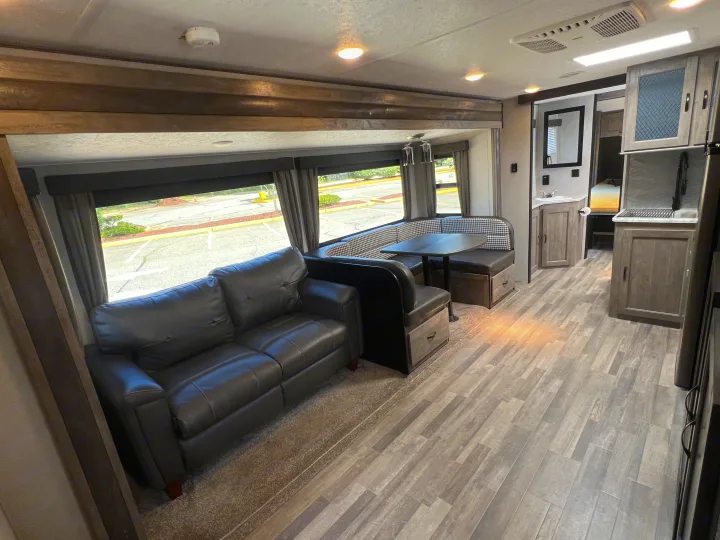 Modern rv rental interior: dark sofa, houndstooth dining booth, kitchenette, wood-look floor, and large windows revealing an outdoor view.. Forest River Vibe 2020