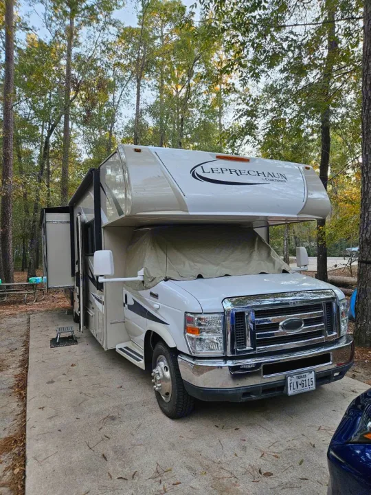 A white Leprechaun RV rental (motorhome) is parked at a treed campsite, its windshield covered.. Forest River Coachmen Leprechaun 2015